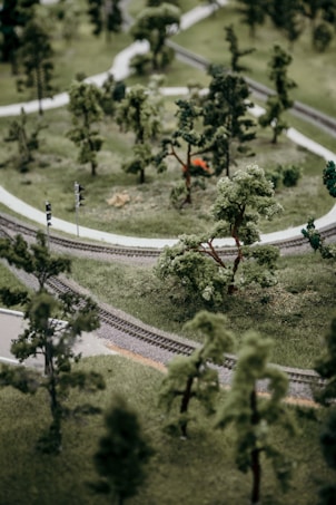 A miniature landscape featuring a winding model railroad track surrounded by lush, green trees and grass. The scene depicts a realistic representation of a natural environment with clear pathways and intricate detailing on the railroad.