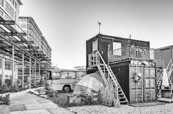 A black and white urban scene featuring a makeshift structure made from shipping containers stacked with a wooden staircase and small balcony. An old bus is parked nearby, with parts of its windows and front obscured by iron beams. Industrial-style buildings are visible in the background, and the ground is a mix of paved path and gravel.