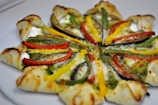A visually appealing pizza with a unique crust design topped with vibrant slices of red, yellow, and green bell peppers, creamy cheese, and a green pesto or sauce. The crust appears to be folded around the edges, creating an interesting pattern.