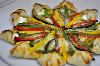 A visually appealing pizza with a unique crust design topped with vibrant slices of red, yellow, and green bell peppers, creamy cheese, and a green pesto or sauce. The crust appears to be folded around the edges, creating an interesting pattern.