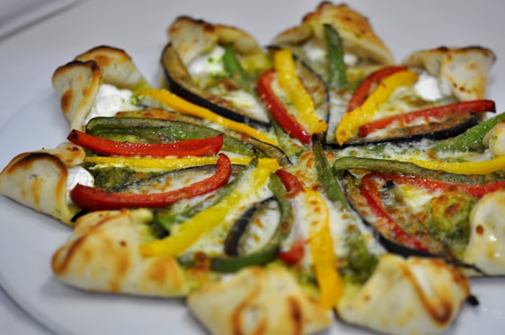 A visually appealing pizza with a unique crust design topped with vibrant slices of red, yellow, and green bell peppers, creamy cheese, and a green pesto or sauce. The crust appears to be folded around the edges, creating an interesting pattern.