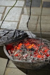 A rustic charcoal grill with burning coals emitting a bright red glow. The grill is placed on a paved surface with its metal lid partially open, allowing a view of the glowing embers and ash.