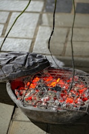 A rustic charcoal grill with burning coals emitting a bright red glow. The grill is placed on a paved surface with its metal lid partially open, allowing a view of the glowing embers and ash.
