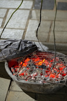 A rustic charcoal grill with burning coals emitting a bright red glow. The grill is placed on a paved surface with its metal lid partially open, allowing a view of the glowing embers and ash.