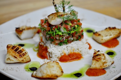 A gourmet dish featuring a central tower of diced vegetables and herbs on a bed of white rice, surrounded by seared scallops and colorful sauces artistically arranged on a white plate. The dish is garnished with a sprig of fresh dill.