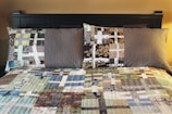 A neatly made bed with two pillows and a patchwork quilt, featuring various geometric patterns and earth tones. The bed frame is dark and adds contrast to the lighter colors of the quilt.