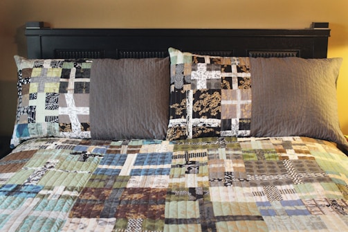 A neatly made bed with two pillows and a patchwork quilt, featuring various geometric patterns and earth tones. The bed frame is dark and adds contrast to the lighter colors of the quilt.