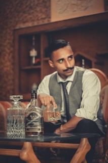 A man with a mustache is seated at a table in a dimly lit room, wearing a white shirt, dark tie, and a vest. He is holding a glass filled with what appears to be whisky and there is a bottle labeled 'Black Dog' and a crystal decanter also on the table. The room features wooden furniture and a textured wall with shelves containing bottles in the background.