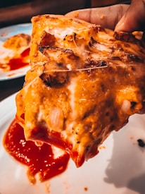 A close-up of a hand holding a slice of pizza with melted cheese and tomato sauce dripping. The pizza appears to have toppings such as onions and possibly chicken, and it is being lifted off a white plate that has some sauce on it.