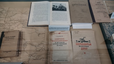 Several old books and documents are laid out on display, including a map and a book open to a black-and-white photograph of several people. The texts appear to be in Russian, suggesting historical or cultural content. The materials seem aged, with some wear and discoloration, hinting at their historical significance.