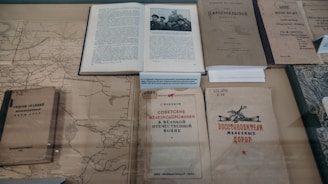 Several old books and documents are laid out on display, including a map and a book open to a black-and-white photograph of several people. The texts appear to be in Russian, suggesting historical or cultural content. The materials seem aged, with some wear and discoloration, hinting at their historical significance.