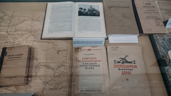 Several old books and documents are laid out on display, including a map and a book open to a black-and-white photograph of several people. The texts appear to be in Russian, suggesting historical or cultural content. The materials seem aged, with some wear and discoloration, hinting at their historical significance.