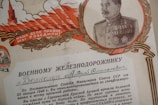 A vintage Soviet Union certificate featuring a detailed illustration of a military officer bordered by a laurel wreath. The officer is dressed in a decorated military uniform with medals. The design includes bold red and golden elements, like feathers and a tower, and text in Cyrillic script.