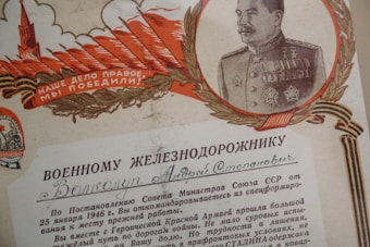 A vintage Soviet Union certificate featuring a detailed illustration of a military officer bordered by a laurel wreath. The officer is dressed in a decorated military uniform with medals. The design includes bold red and golden elements, like feathers and a tower, and text in Cyrillic script.