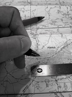 A monochrome image depicting a close-up of a hand holding a pen over a map. A ruler is also placed on the map, indicating measurements or planning activity. The map details roads and locations, with 'Paris' marked prominently.