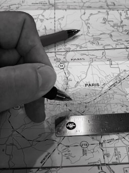 A monochrome image depicting a close-up of a hand holding a pen over a map. A ruler is also placed on the map, indicating measurements or planning activity. The map details roads and locations, with 'Paris' marked prominently.