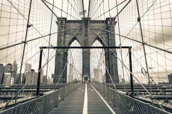 This is an image of a bridge with a strong mesh of cables suspending its structure. The bridge has a prominent stone archway in the center and a distinct pedestrian pathway with a thick white line in the middle. The background reveals a city skyline with towering buildings, and the sky is overcast with clouds.