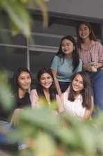 Five young women are sitting together, smiling and appearing relaxed. They are positioned closely, suggesting a sense of friendship or camaraderie. The focus is on them, with some greenery blurred in the foreground, adding a natural element to the composition. The setting appears to be casual or informal.