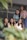 Five young women are sitting together, smiling and appearing relaxed. They are positioned closely, suggesting a sense of friendship or camaraderie. The focus is on them, with some greenery blurred in the foreground, adding a natural element to the composition. The setting appears to be casual or informal.
