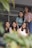 Five young women are sitting together, smiling and appearing relaxed. They are positioned closely, suggesting a sense of friendship or camaraderie. The focus is on them, with some greenery blurred in the foreground, adding a natural element to the composition. The setting appears to be casual or informal.