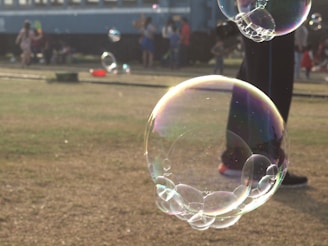 A large, translucent bubble floats in the air with several smaller bubbles attached to its surface. In the background, there is a blurred image of a group of people, possibly children, on a grassy area near a train or bus. The sunlight creates colorful reflections on the bubbles, adding to their visual appeal.