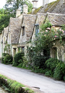A row of charming stone cottages with steeply pitched roofs covered in moss. The cottages are adorned with lush greenery and blooming flowers, while a narrow path runs alongside them.