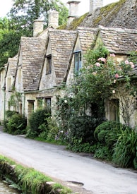 A row of charming stone cottages with steeply pitched roofs covered in moss. The cottages are adorned with lush greenery and blooming flowers, while a narrow path runs alongside them.
