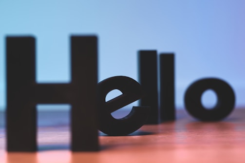 Large black 3D letters on a wooden surface spelling out the word 'Hello'. The background transitions from light blue to purple, creating a soft gradient effect.