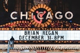 A brightly lit marquee advertising a performance by Brian Regan on December 31 at 8 PM. The marquee is part of a larger theater sign, adorned with numerous lights and a large, illuminated 'Chicago' sign at the top. A person is visible standing on a raised platform in front of the marquee.