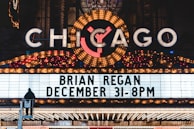 A brightly lit marquee advertising a performance by Brian Regan on December 31 at 8 PM. The marquee is part of a larger theater sign, adorned with numerous lights and a large, illuminated 'Chicago' sign at the top. A person is visible standing on a raised platform in front of the marquee.