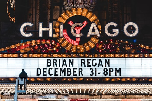 A brightly lit marquee advertising a performance by Brian Regan on December 31 at 8 PM. The marquee is part of a larger theater sign, adorned with numerous lights and a large, illuminated 'Chicago' sign at the top. A person is visible standing on a raised platform in front of the marquee.