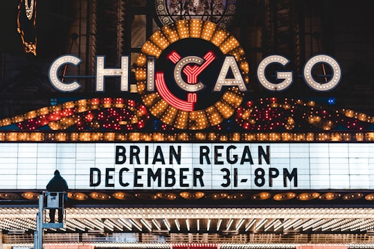 A brightly lit marquee advertising a performance by Brian Regan on December 31 at 8 PM. The marquee is part of a larger theater sign, adorned with numerous lights and a large, illuminated 'Chicago' sign at the top. A person is visible standing on a raised platform in front of the marquee.