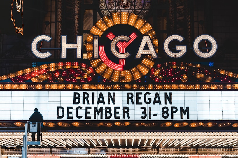 A brightly lit marquee advertising a performance by Brian Regan on December 31 at 8 PM. The marquee is part of a larger theater sign, adorned with numerous lights and a large, illuminated 'Chicago' sign at the top. A person is visible standing on a raised platform in front of the marquee.