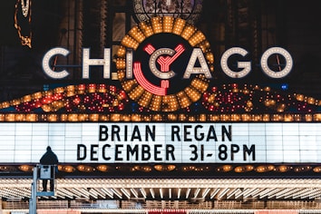A brightly lit marquee advertising a performance by Brian Regan on December 31 at 8 PM. The marquee is part of a larger theater sign, adorned with numerous lights and a large, illuminated 'Chicago' sign at the top. A person is visible standing on a raised platform in front of the marquee.