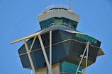 A modern airport control tower with a sleek, angular design featuring tinted windows and structural columns. The sky in the background is clear and blue, adding a vibrant contrast to the neutral tones of the building.