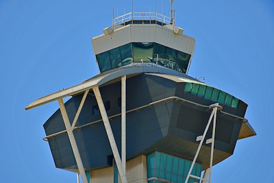 A modern airport control tower with a sleek, angular design featuring tinted windows and structural columns. The sky in the background is clear and blue, adding a vibrant contrast to the neutral tones of the building.