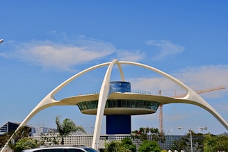 A futuristic building with a circular design supported by four large, curved arches. It features a central blue cylindrical structure with windows on the top level, and it is surrounded by lush greenery and a few cars in the foreground. There are cranes and a clear blue sky in the background.