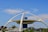 A futuristic building with a circular design supported by four large, curved arches. It features a central blue cylindrical structure with windows on the top level, and it is surrounded by lush greenery and a few cars in the foreground. There are cranes and a clear blue sky in the background.