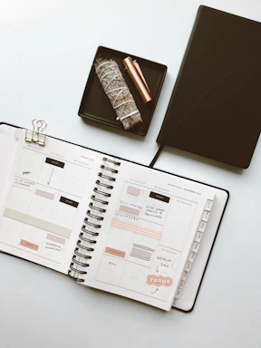 A well-organized flat lay featuring an open planner filled with handwritten notes and stickers. Next to the planner is a black notebook, a decorative bundle of sage, and a bronze-colored pen, all neatly arranged on a small black tray.
