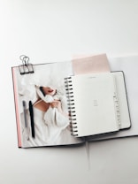 An open planner is laid out on a table, featuring a page with the text 'You got this'. Beside it, there is a notebook with a magazine partially visible, showing a person in a white garment. Two pens are placed on top of the magazine, and a binder clip is attached to the notebook.