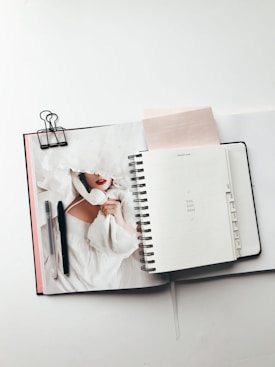 An open planner is laid out on a table, featuring a page with the text 'You got this'. Beside it, there is a notebook with a magazine partially visible, showing a person in a white garment. Two pens are placed on top of the magazine, and a binder clip is attached to the notebook.