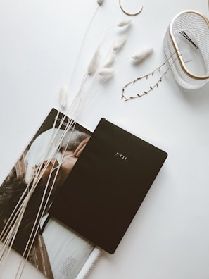A minimalist arrangement on a white surface features a black planner labeled 'STIL' placed on top of an open magazine. Beside it, there are decorative elements, including fluffy white dried grasses and a small container holding a few hairpins. The overall aesthetic is clean and elegant.