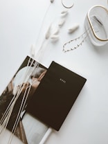A minimalist arrangement on a white surface features a black planner labeled 'STIL' placed on top of an open magazine. Beside it, there are decorative elements, including fluffy white dried grasses and a small container holding a few hairpins. The overall aesthetic is clean and elegant.