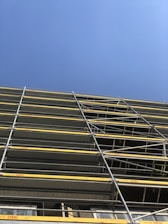 A construction site featuring a tall scaffolding structure with multiple levels of metal framework and yellow safety barriers against a clear blue sky.
