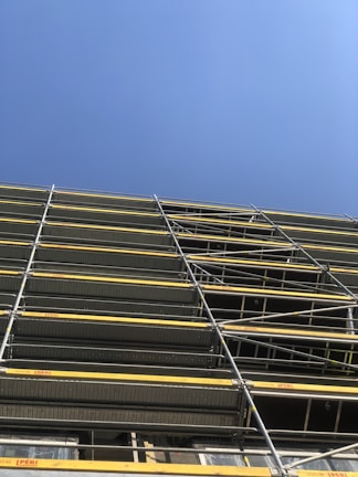 A construction site featuring a tall scaffolding structure with multiple levels of metal framework and yellow safety barriers against a clear blue sky.