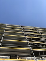 A construction site featuring a tall scaffolding structure with multiple levels of metal framework and yellow safety barriers against a clear blue sky.