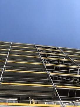 A construction site featuring a tall scaffolding structure with multiple levels of metal framework and yellow safety barriers against a clear blue sky.