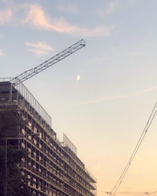 A construction site features a large crane against a clear sky with a visible crescent moon. The building under construction has scaffolding along its side, and there are visible power lines cutting across the scene. The sky is tinged with soft hues of pink and blue, indicating either dawn or dusk.