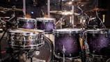 A drum set with sparkling dark purple shells, surrounded by various cymbals and microphones, is featured in a stage setting. The lighting reflects off the shiny surfaces, creating a vibrant and lively atmosphere.