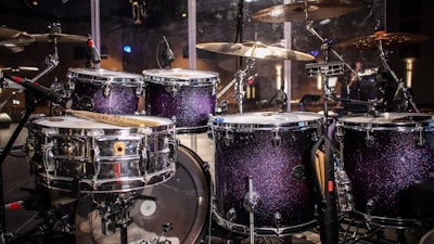 A drum set with sparkling dark purple shells, surrounded by various cymbals and microphones, is featured in a stage setting. The lighting reflects off the shiny surfaces, creating a vibrant and lively atmosphere.